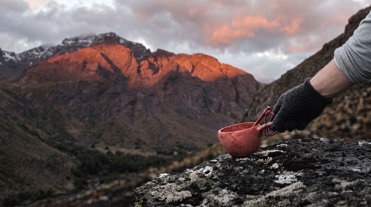 Mano con guante deja una taza Kupilka roja sobre una roca, con la cordillera al atardecer